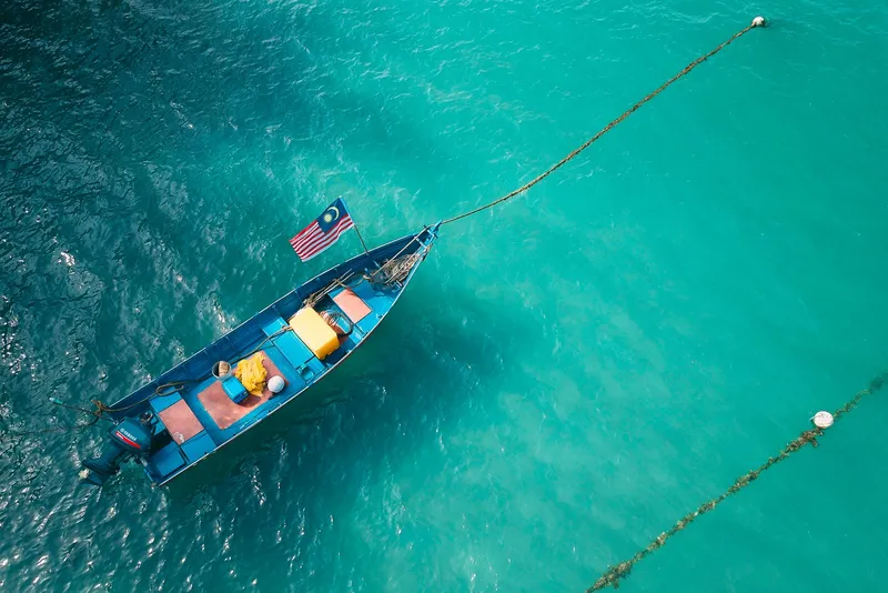 Small blue boat flying a Malaysian flag, seen from above on the sea