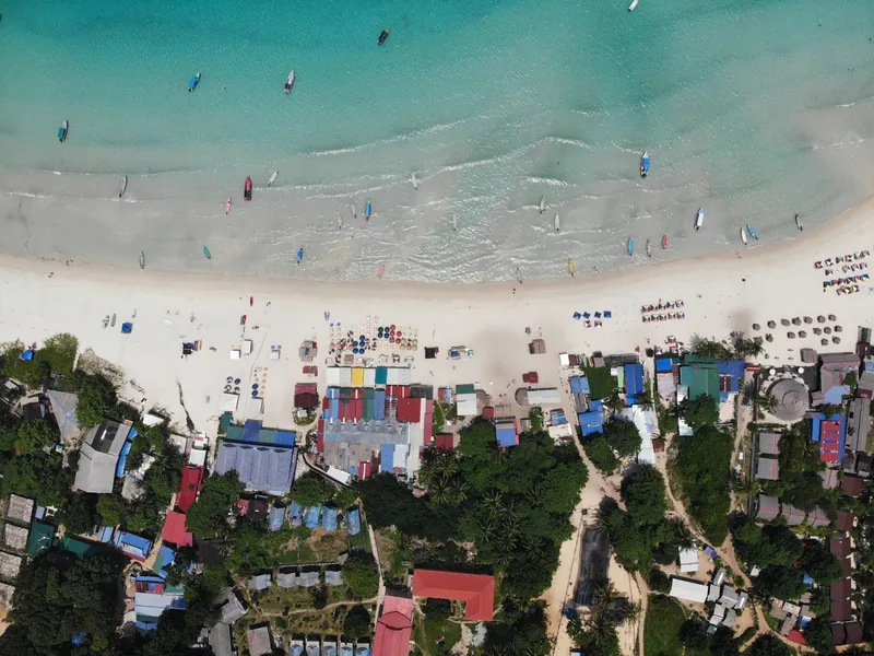Drone view of boats near a sandy beach, with houses and green trees on the shore