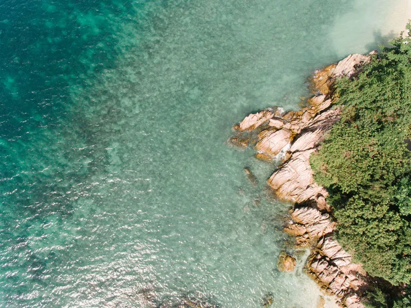 Drone view of turquoise sea along a rocky, tree-lined coast
