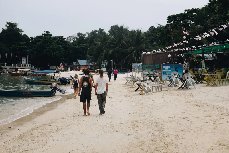 Two people walking on a sandy beach, boats and beachfront seating behind